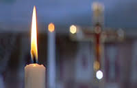Vitt ljus brinner med stor låga, i bakgrunden syns konturerna av ett kors.